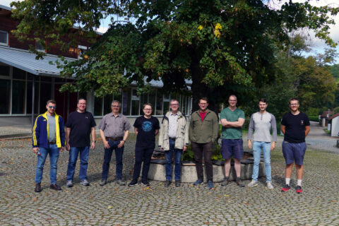 We can see a group photo. Nine men stand next to each other. There are two professors and seven employees. The men stand in front of a large tree and look at the camera with a relaxed smile. It is late summer.