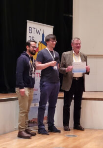 The photo shows three men on stage. On the left we see two organizers of the demo session, and on the right is Prof. Meyer-Wegener. He has just received the Best Demo Award from the organizers and is proudly holding it up to the camera. In the background there is a floor display with the BTW25 conference logo.